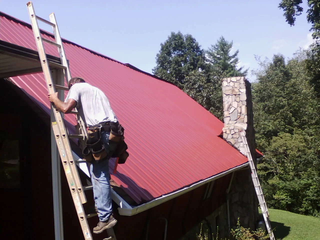 Roofer on a ladder beside a red metal roof and stone chimney — Soffit & Fascia Repair by Roofing Brothers in Millville